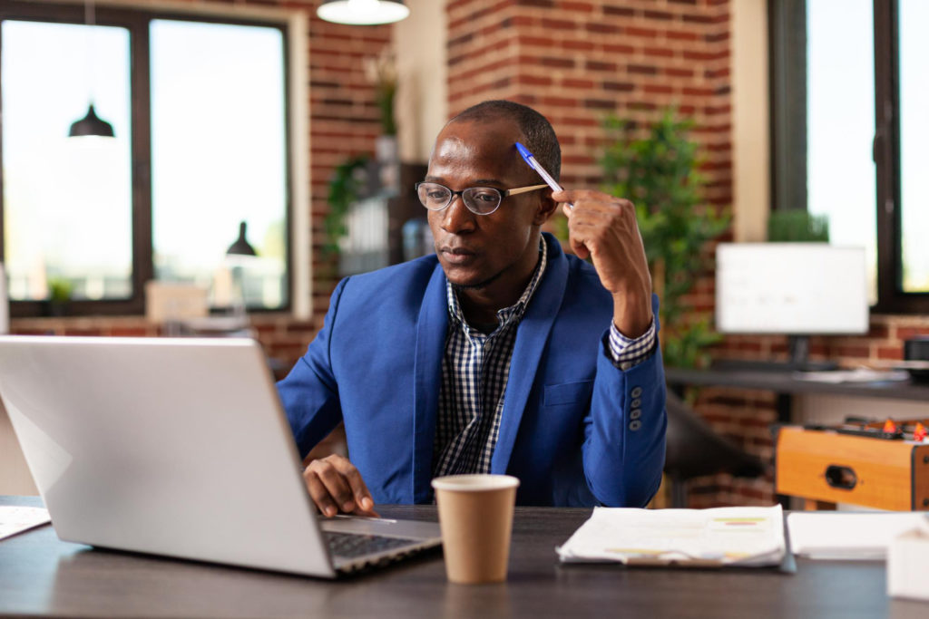 Um homem negro, vestindo terno azul, está em frente a uma mesa de escritório marrom. Ele olha para um laptop prata e apoia uma caneta de tampa azul em sua cabeça, como se estivesse pensando. 