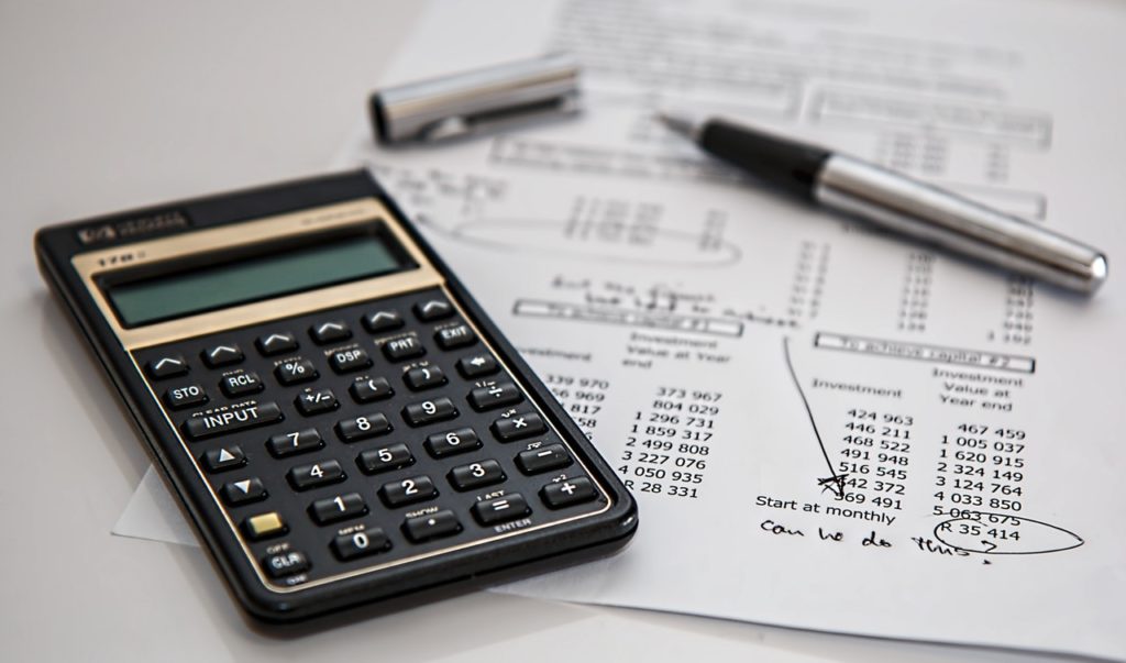 Uma calculadora preta está em cima de uma folha branco com dados de custos. Ambos estão sobre uma mesa branca e representam o cálculo necessário para fazer a redução de custos.