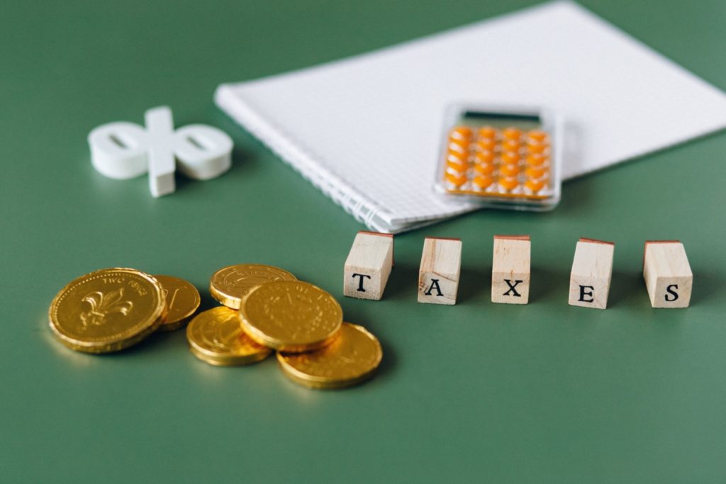 Algumas moedas douradas, letras de madeira que formam a palavra “taxes”, um símbolo de porcentagem, uma calculadora e um caderno estão sobre uma mesa verde. 