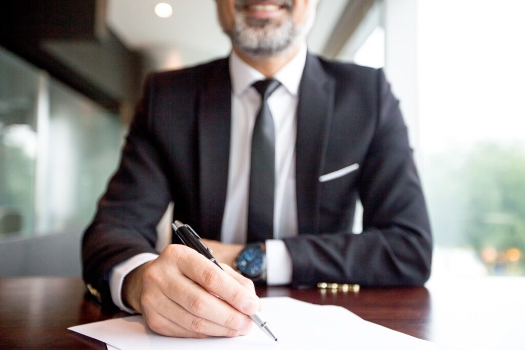 Um homem branco, de barba por fazer, usando terno preto e um relógio de pulso, está sentado e sorrindo, enquanto usa uma caneta para escrever em folhas de papel.